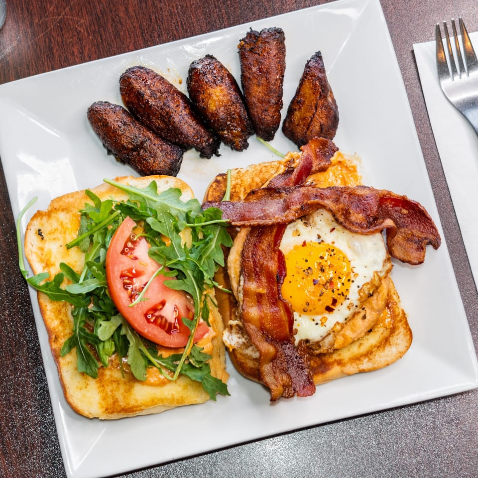 A breakfast plate with French toast, bacon, fried egg, tomato, arugula, and plantains.