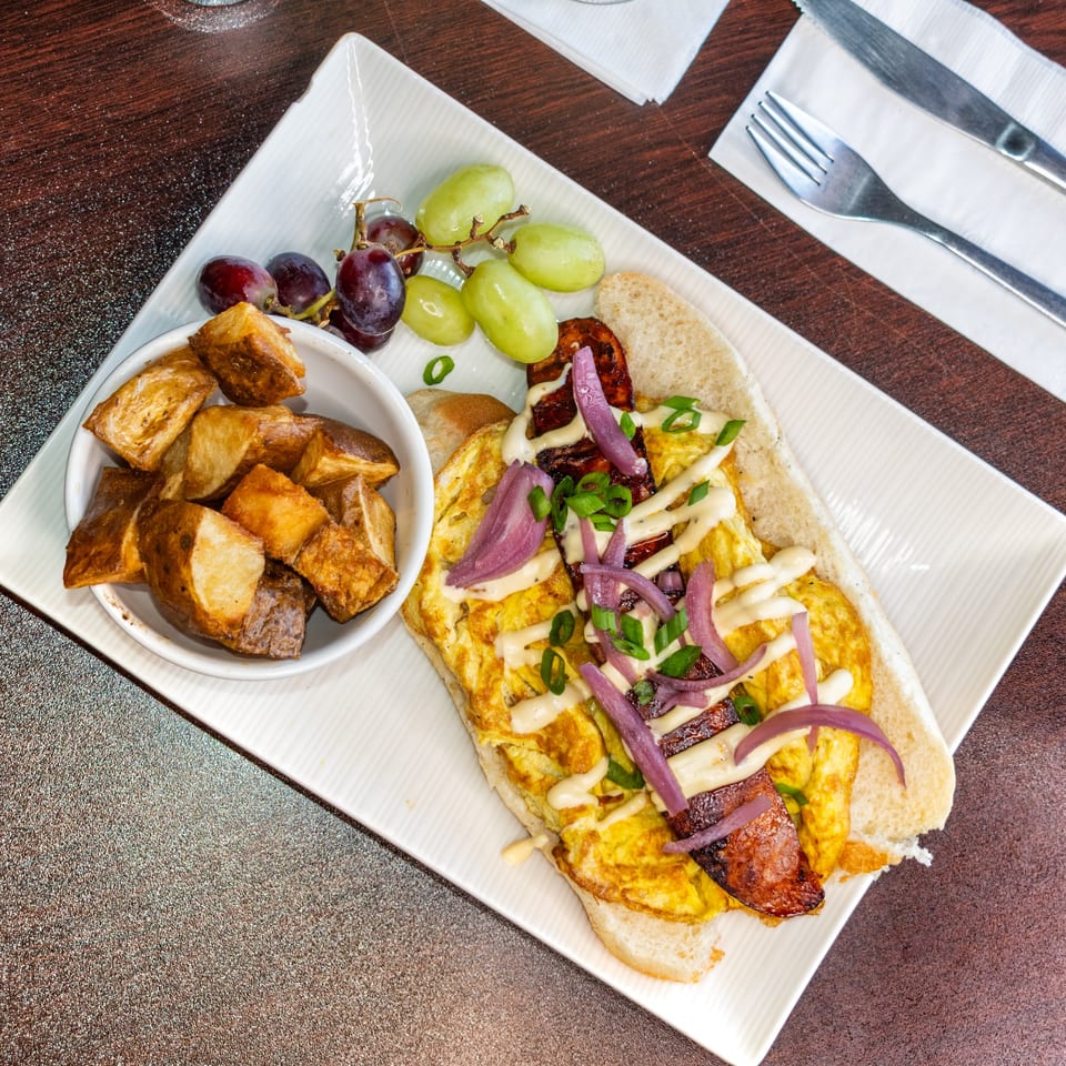 Breakfast sandwich with eggs, bacon, red onion, and sauce, served with roasted potatoes and grapes.