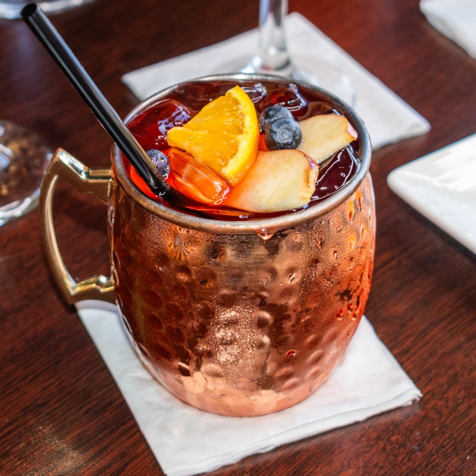 A copper mug filled with a dark red drink, ice, orange slice, blueberries, and apple pieces.