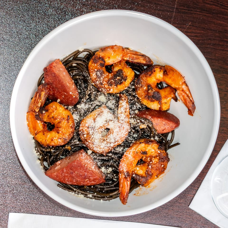 A white bowl of black spaghetti topped with grilled shrimp, sausage, and parmesan cheese.